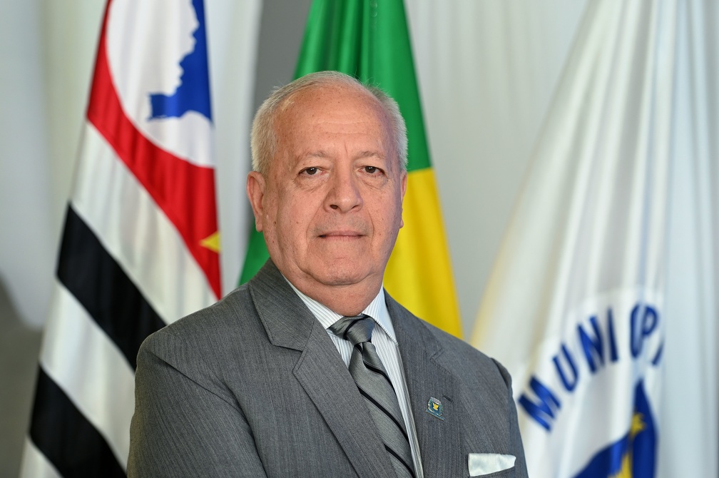 Fotografia do secretário Arly de Lara Romeo, de pé, com bandeiras ao fundo