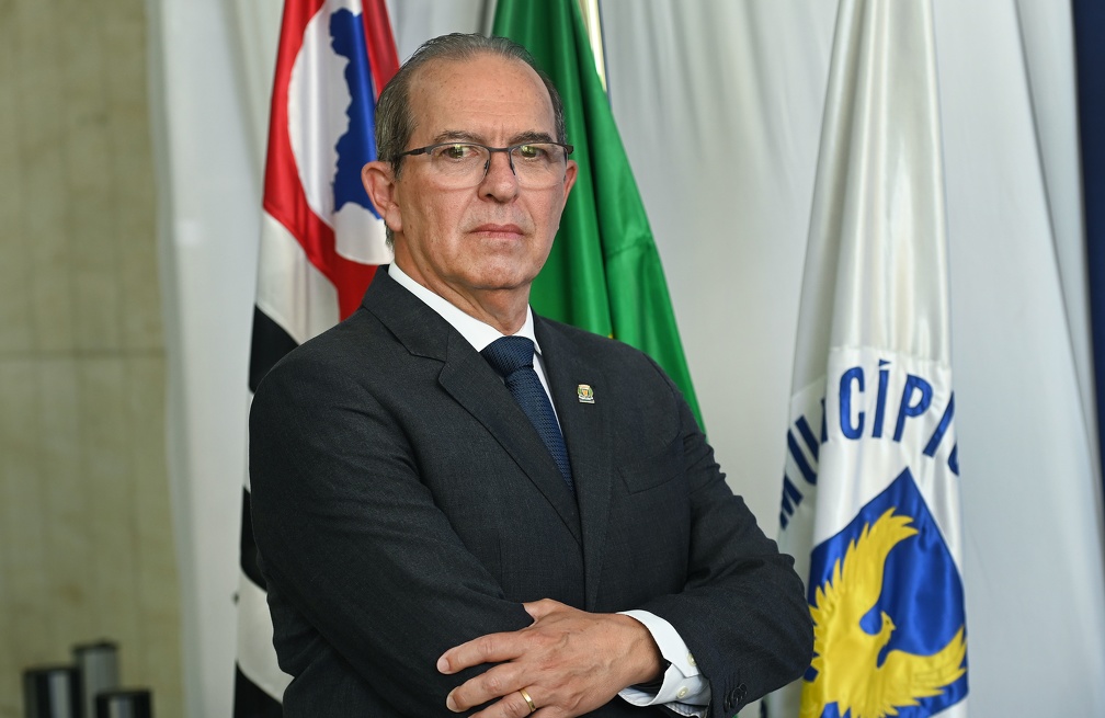 Fotografia do secretário Aurílio Sérgio Costa Caiado, sentado, com as bandeiras de Campinas, do Estado de São Paulo e do Brasil ao fundo