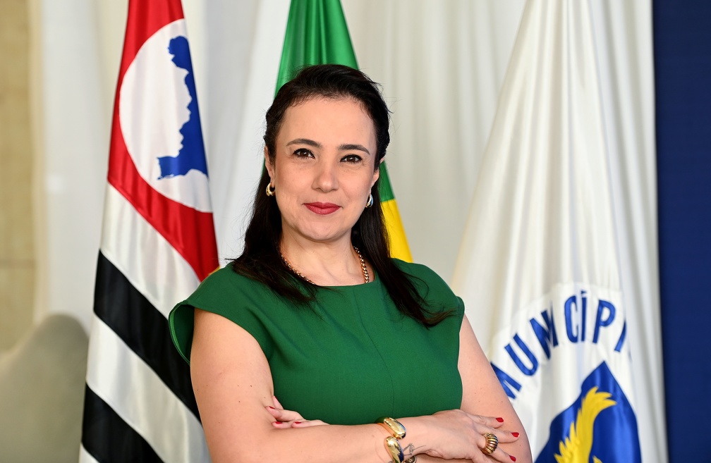 Fotografia da secretária Carolina Baracat Lazinho, sorrindo, com bandeiras ao fundo