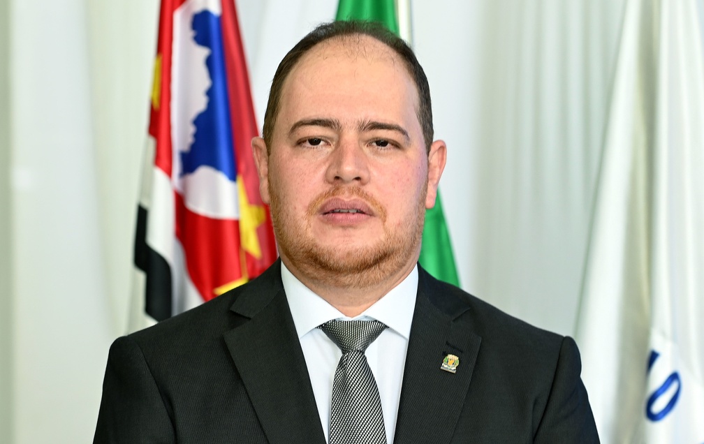 Fotografia do secretário Christiano Biggi Dias, sentado, com as bandeiras de Campinas, do Estado de São Paulo e do Brasil ao fundo