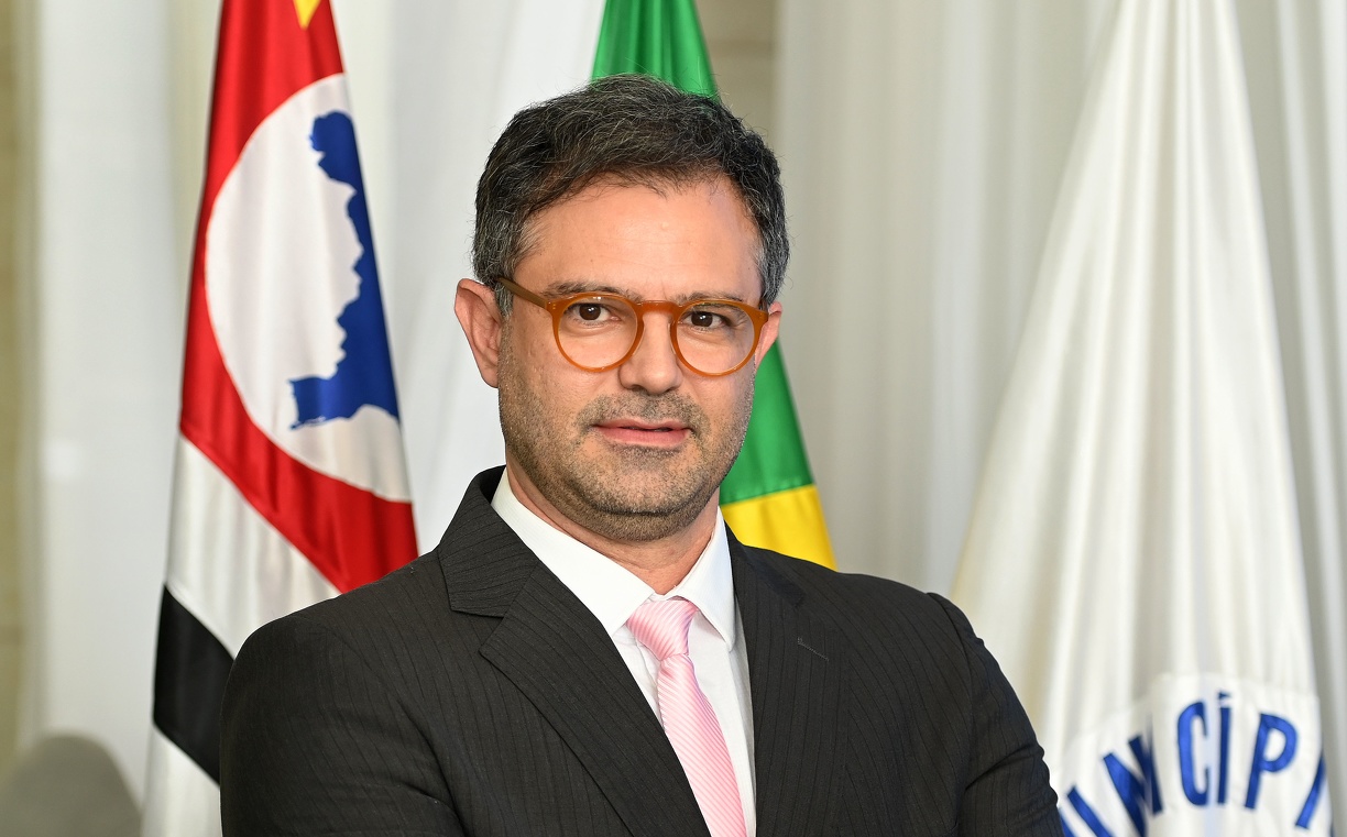 Fotografia do secretário Fernando Vanin, de pé, com bandeiras ao fundo