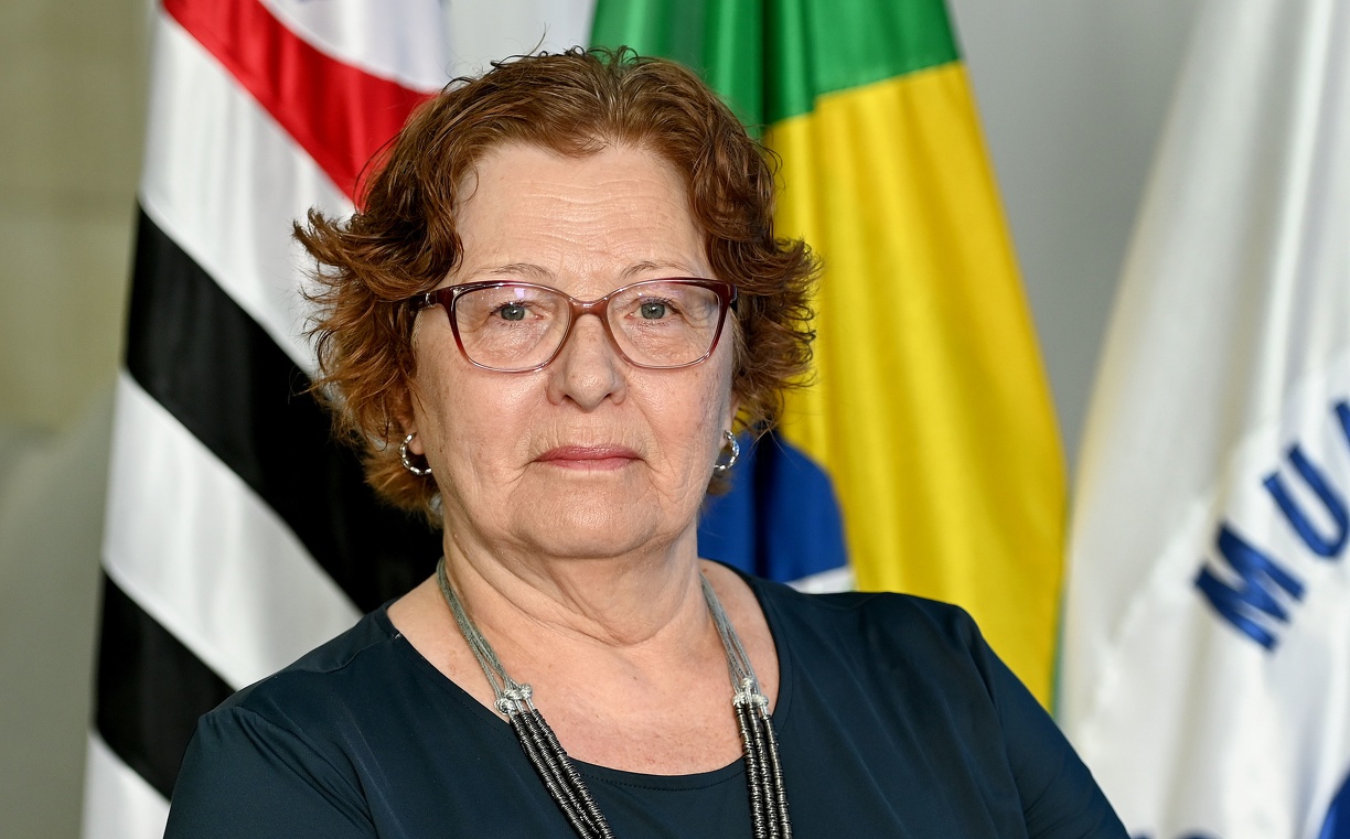 Fotografia da secretária Maria Emilia de Arruda Faccioni, com as bandeiras de Campinas, do Brasil e do Estado de São Paulo ao fundo