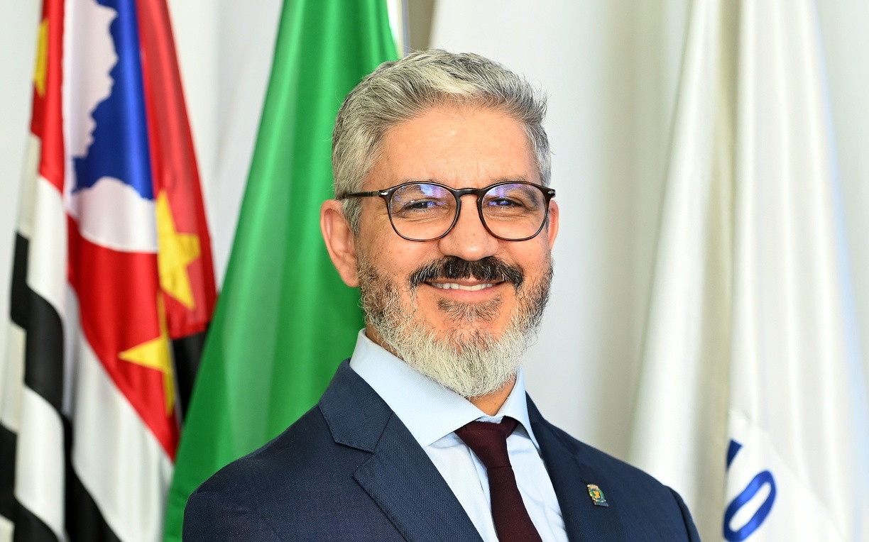 Fotografia do secretário Peter Panutto Panont, em pé, com as bandeiras de Campinas, do Estado de São Paulo e do Brasil ao fundo