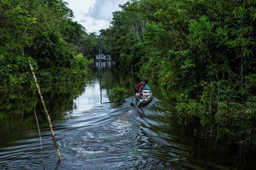 Morador conduz barco por igarapé, braço estreito de rio que corta a floresta, no caminho de casa erguida sobre palafitas; o cotidiano ribeirinho é um dos temas da exposição