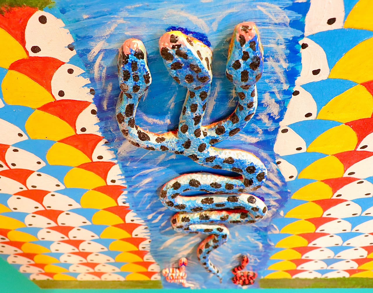 Escultura de serpente entalhada e pintada em azul e branco, com manchas pretas, montada sobre tela com padrão de escamas coloridas