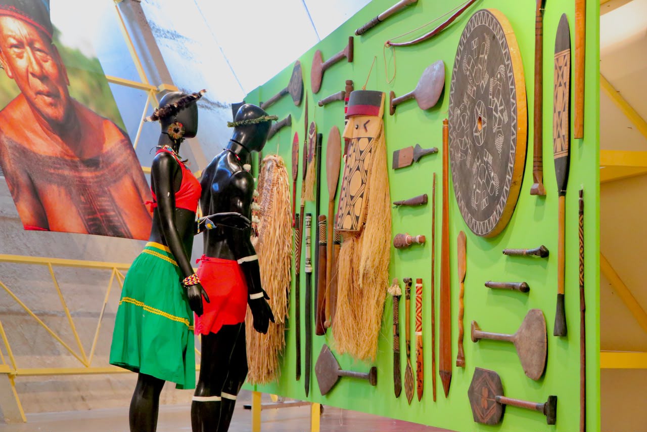Painel expositivo reúne utensílios e armas indígenas: remos, bordunas, escudos e outros objetos entalhados, ao lado de manequins com trajes cerimoniais