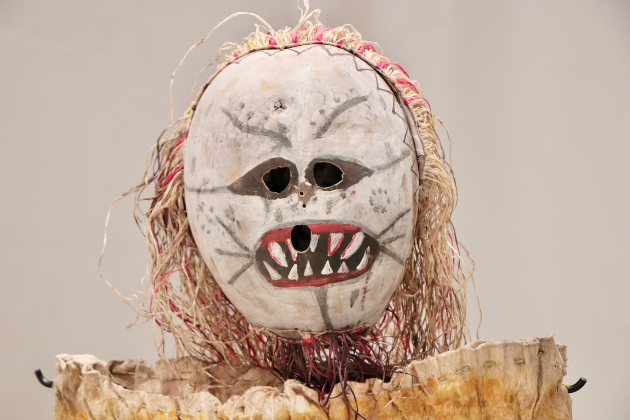 Máscara ritual indígena, com base branca, olhos vazados e boca pintada com dentes, adornada com fibras naturais